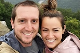 A young man and woman stand grinning while taking a selfie on top of a misty mountain