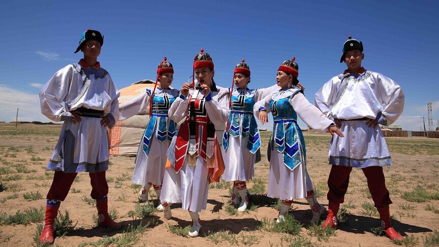 Six Mongolian performers dressed in white robes and wearing hats pose at Naadam Festival.