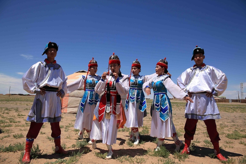 Six Mongolian performers dressed in white robes and wearing hats pose at Naadam Festival.