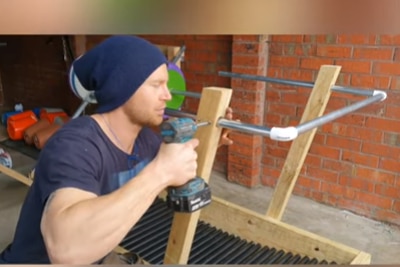 A man with a beanie drills a metal pole on wood to make a conveyor belt. 