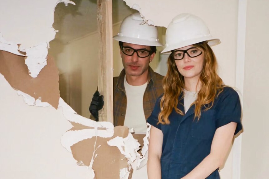 Emma and Nathan stand on either side of a knocked-through wall wearing hard hats, Emma with a hammer in hand, looking smug