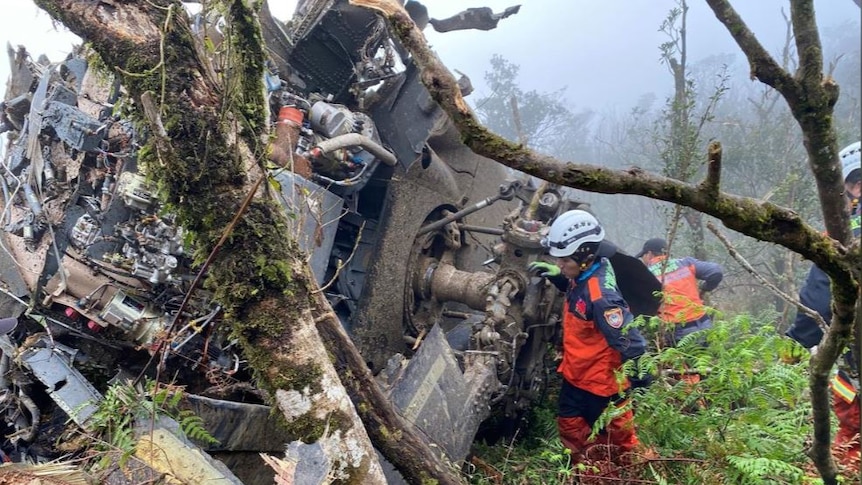 The twisted wreckage of a Black Hawk helicopter in thick foggy forest with two rescuers near it.