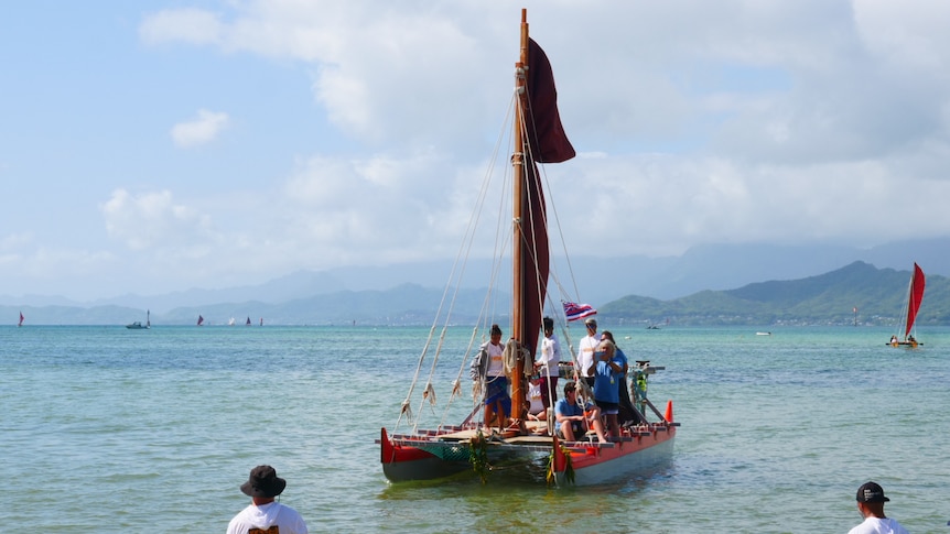A group of people standing on a traditional canoe sail towards shore.