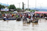 A crowd of people stand at a waterfront behind small boats bobbing on high waters.