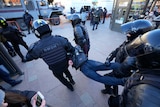 Riot police carry a man away, with two police holding him at his shoulders, and two other lifting his feet. 