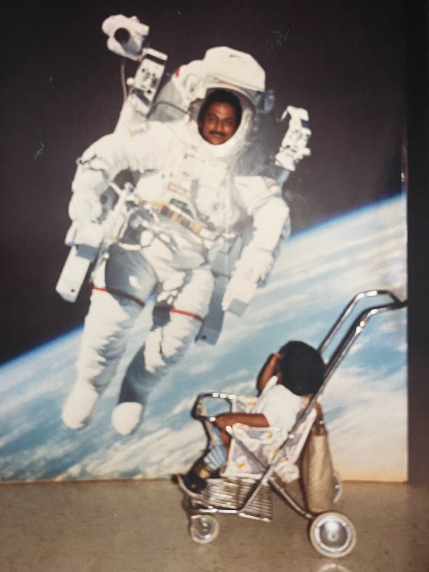 A young child sitting in a pram looks up as a man standing behind a cut-out of an astronaut.