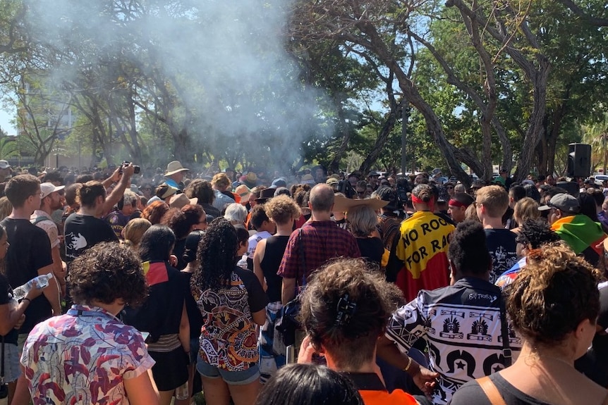 A large crowd of people with Aboriginal and Torres Strait Islander flags gather in a park.