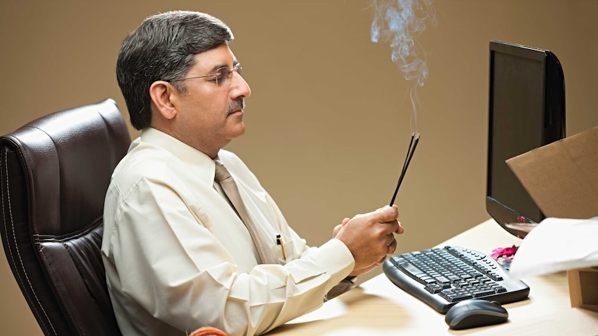 Indian man sits and office desk in front of computer, holding incense and closing his eyes.