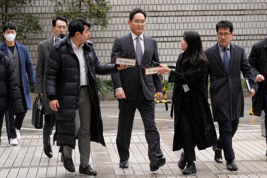 Four men in suits and one woman in a long coat walk on tiles. One woman and one man are holding a box with tiny microphones.
