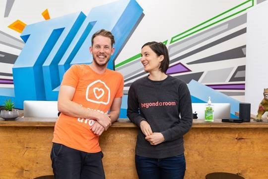 A man and woman in t-shirts lean on a desk in an office with a brightly coloured mural painted on the wall.