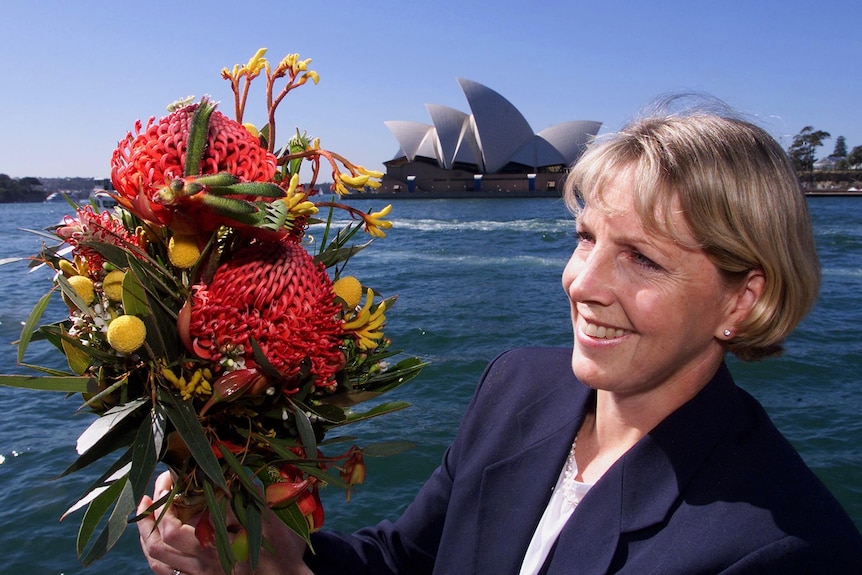 A woman smiles at the camera as she holds a floral bouquet with the Sydney Opera House in the background. 