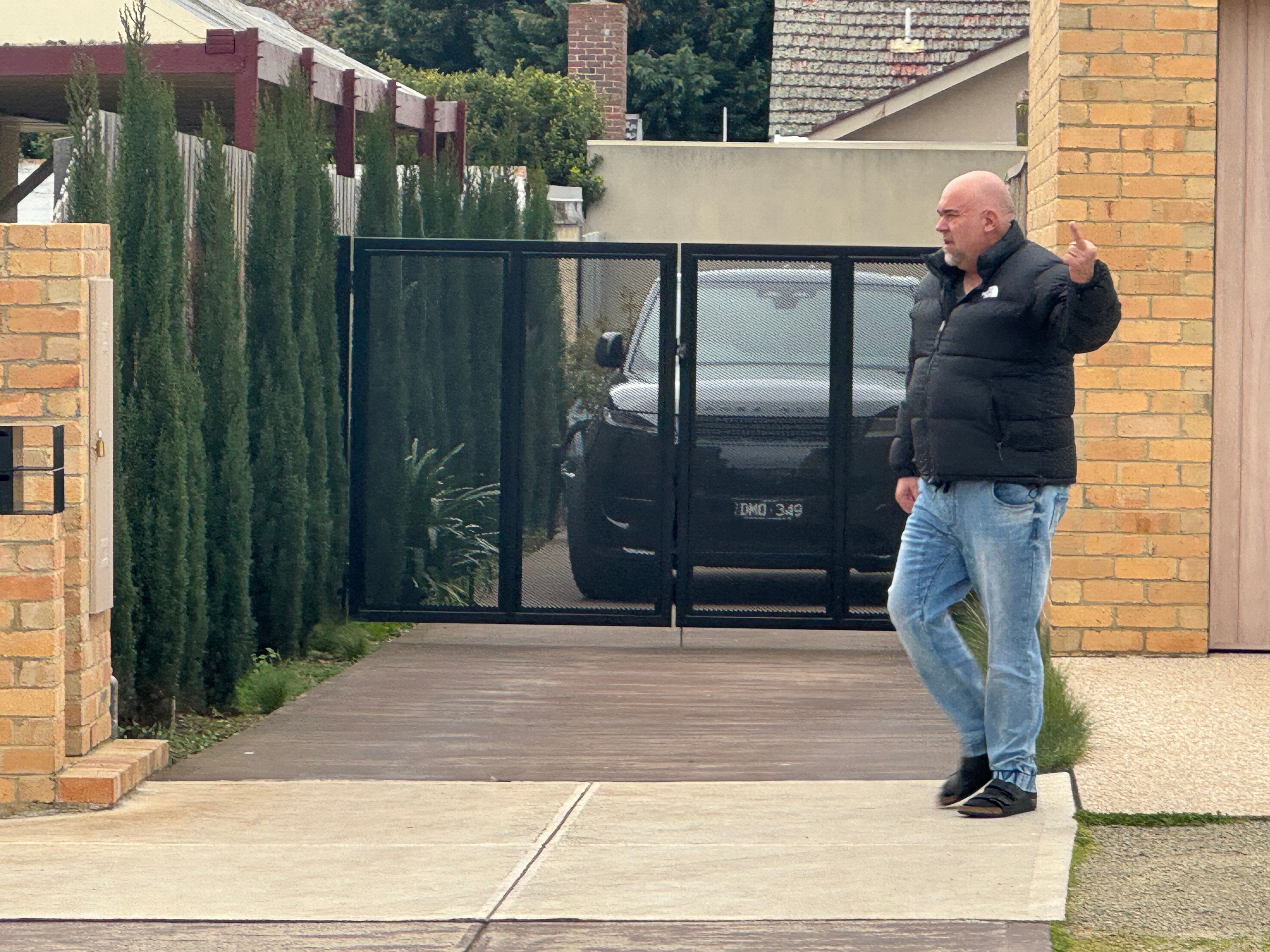 A man gestures toward the camera while walking across a drive way in front of a black SUV.