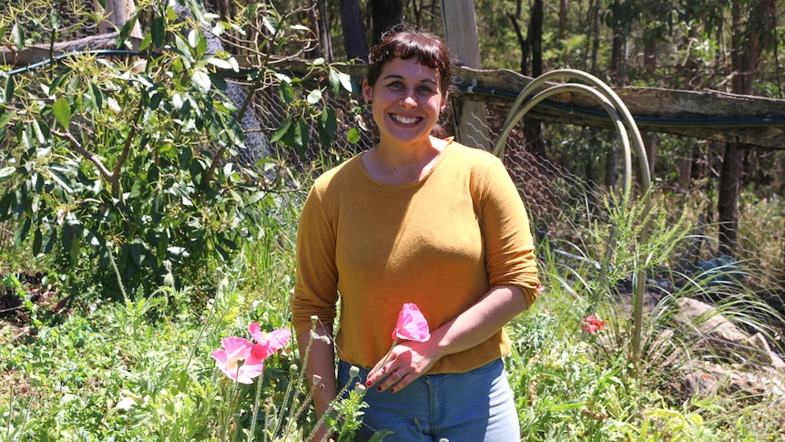 A lady in an orange jumper and blue jeans holds onto pink flowers in a garden while smiling at the camera.