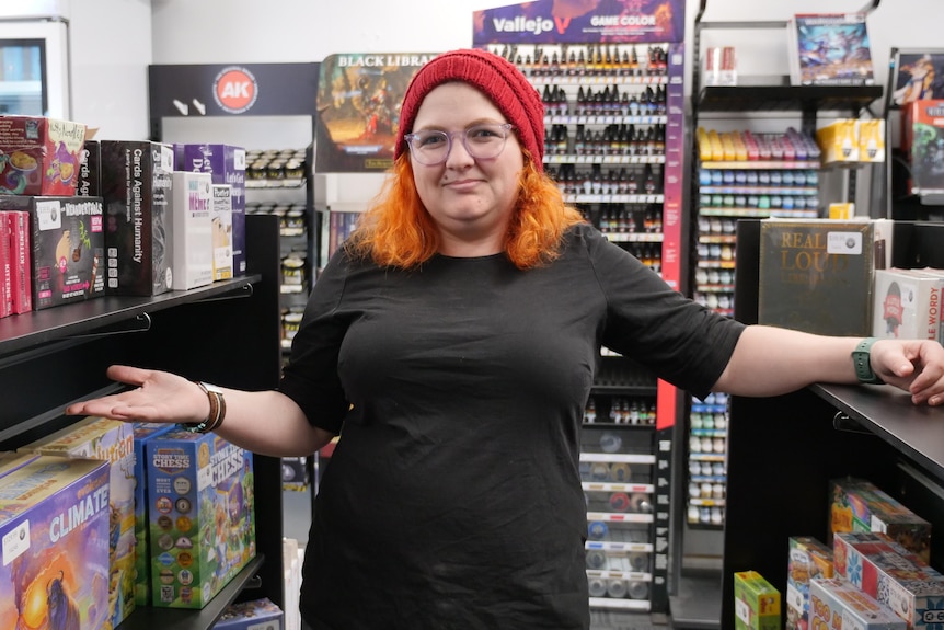 Woman wearing all black with orange hair and red beanie gestures towards boardgames on a shelf