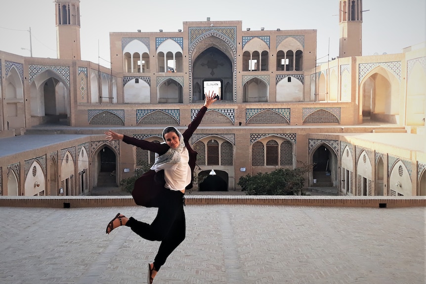 Woman wearing head scarf jumps for joy in front of Iranian building