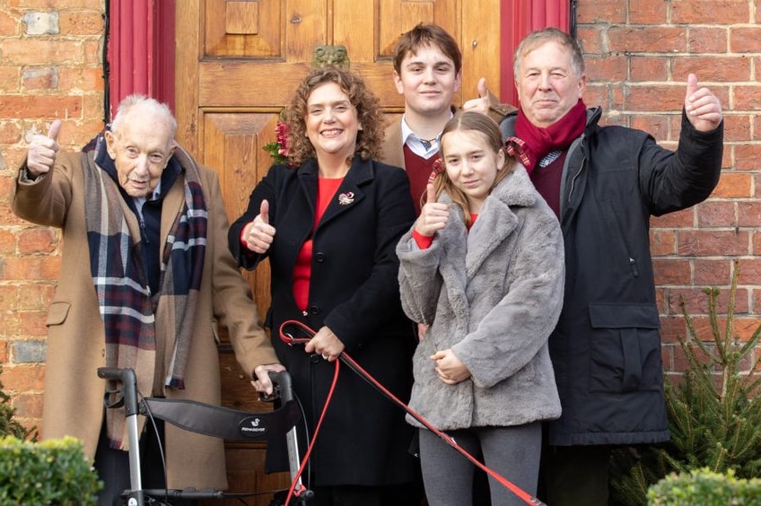 White family in coats stands outside brick house with red door