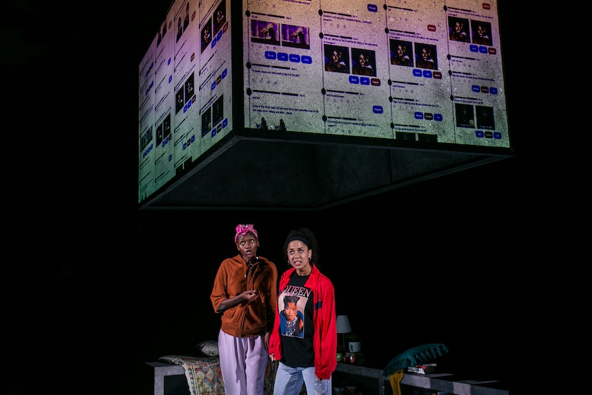 Two women stand under a large square-shaped screen on a stage. Tweets are projected on the screen.