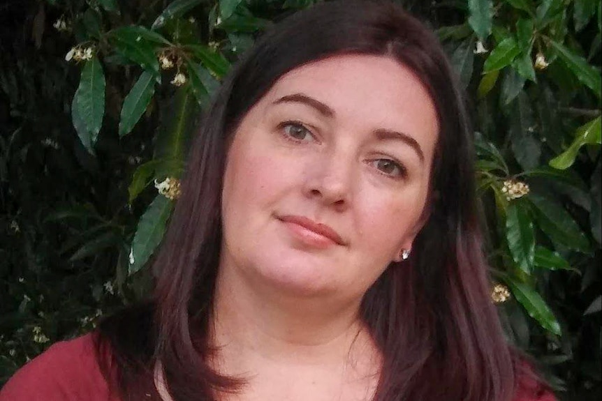 A young woman with dark hair stands in front of a green tree