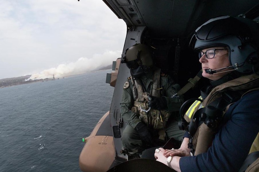 a woman with glasses and a helmet on sits next to a person in full military uniform as she looks out a helicopter to the sea