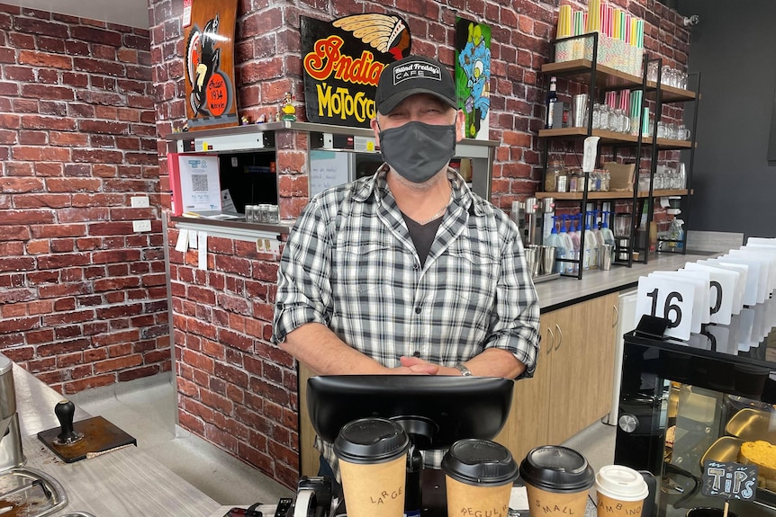 Man at cash register with coffee cups at the front. He is wearing a mask and cafe is empty