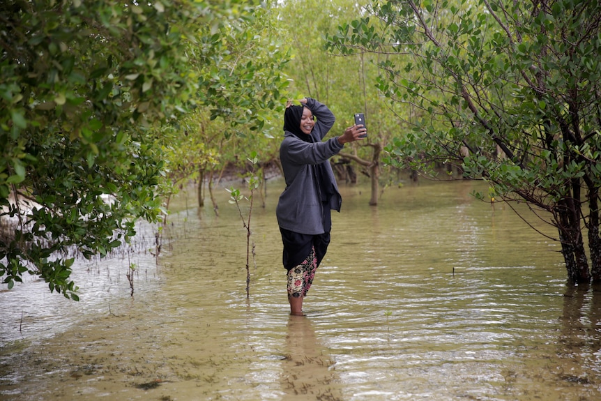 A woman stands in front of mangrove trees and takes a selfie.