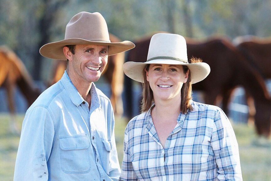 A man wearing jeans, a blue shirt and brown cowboy hat stands next to a woman in jeans, a blue and white checked shirt and hat 