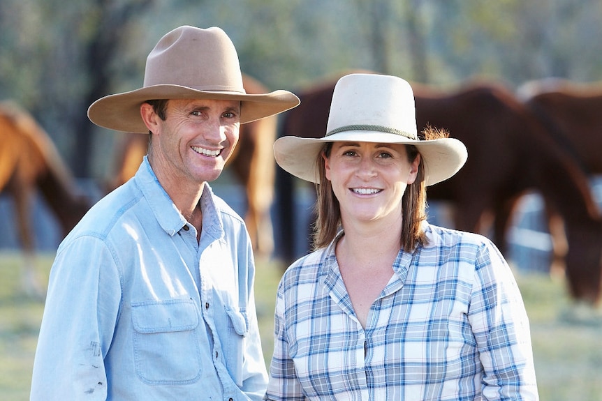 A man wearing jeans, a blue shirt and brown cowboy hat stands next to a woman in jeans, a blue and white checked shirt and hat 