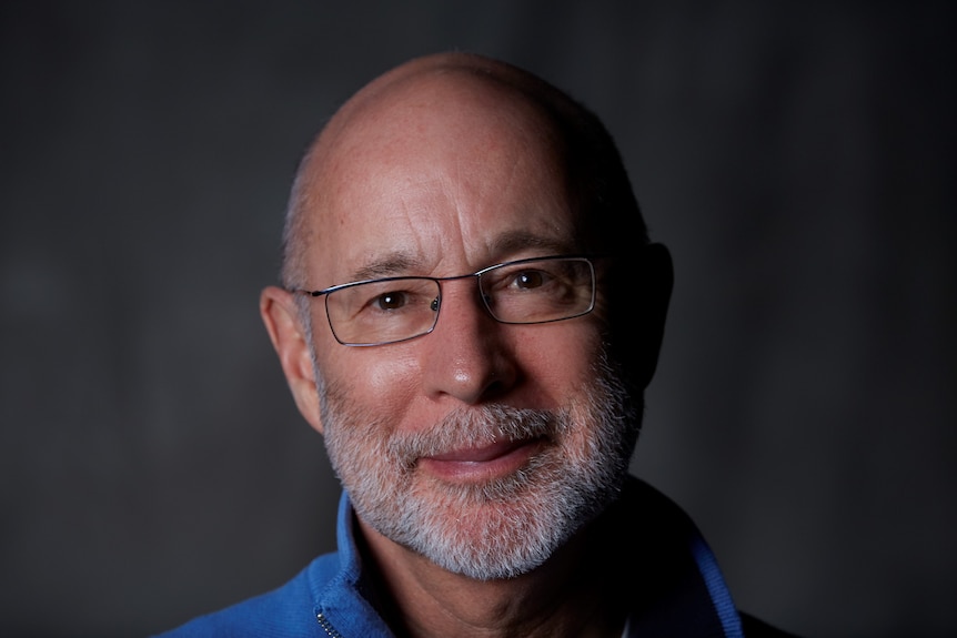 Man smiles at camera in close up portrait, wears blue shirt and rectangular shaped steel rimmed glasses