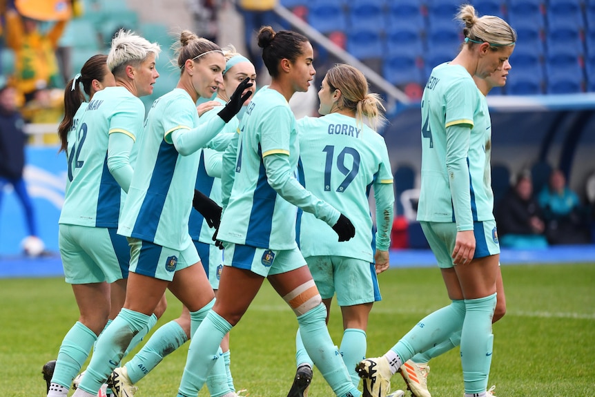 matildas soccer players on the field in their aqua coloured jersey and shorts celebrating a goal