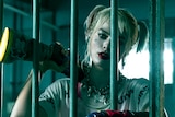 A woman with pale make-up and pigtails, stands behind the bars of a jail cell and rests a long firearm on one shoulder.