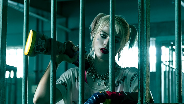 A woman with pale make-up and pigtails, stands behind the bars of a jail cell and rests a long firearm on one shoulder.