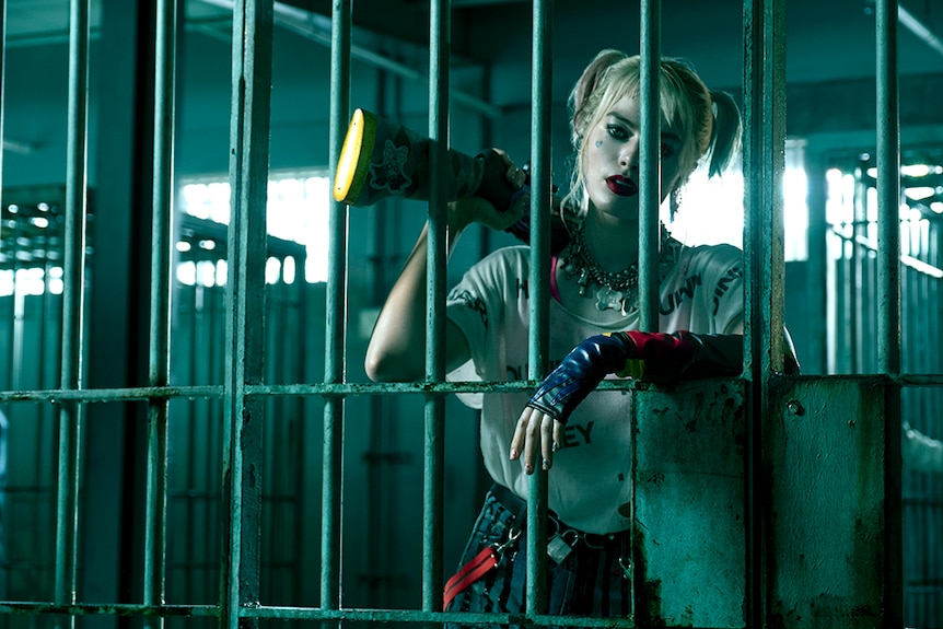 A woman with pale make-up and pigtails, stands behind the bars of a jail cell and rests a long firearm on one shoulder.