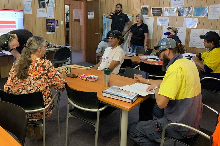 A group of people are sitting around desks in a classroom. There is a large computer screen in the background.