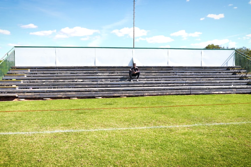 An oval with green grass and stadium seating. There is a young man sitting on the seats alone. The sky is blue above.