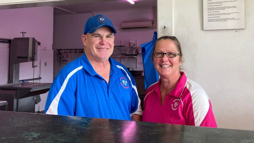 Butcher and his wife stand smiling behind counter of butchery. 