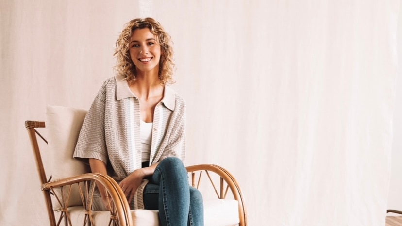 A woman with curly blonde hair wearing jeans and a beige shirt sits on a chair, smiling to the camera.