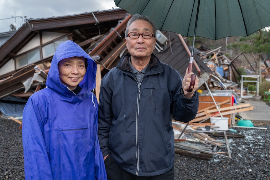 On the left a woman in a blue raincoat, next to her is a man holding an umbrella. Behind them is debris.