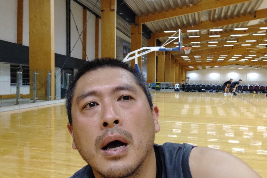 Selfie of Christian looking tired at an indoor basketball court, with a game in play in the background.