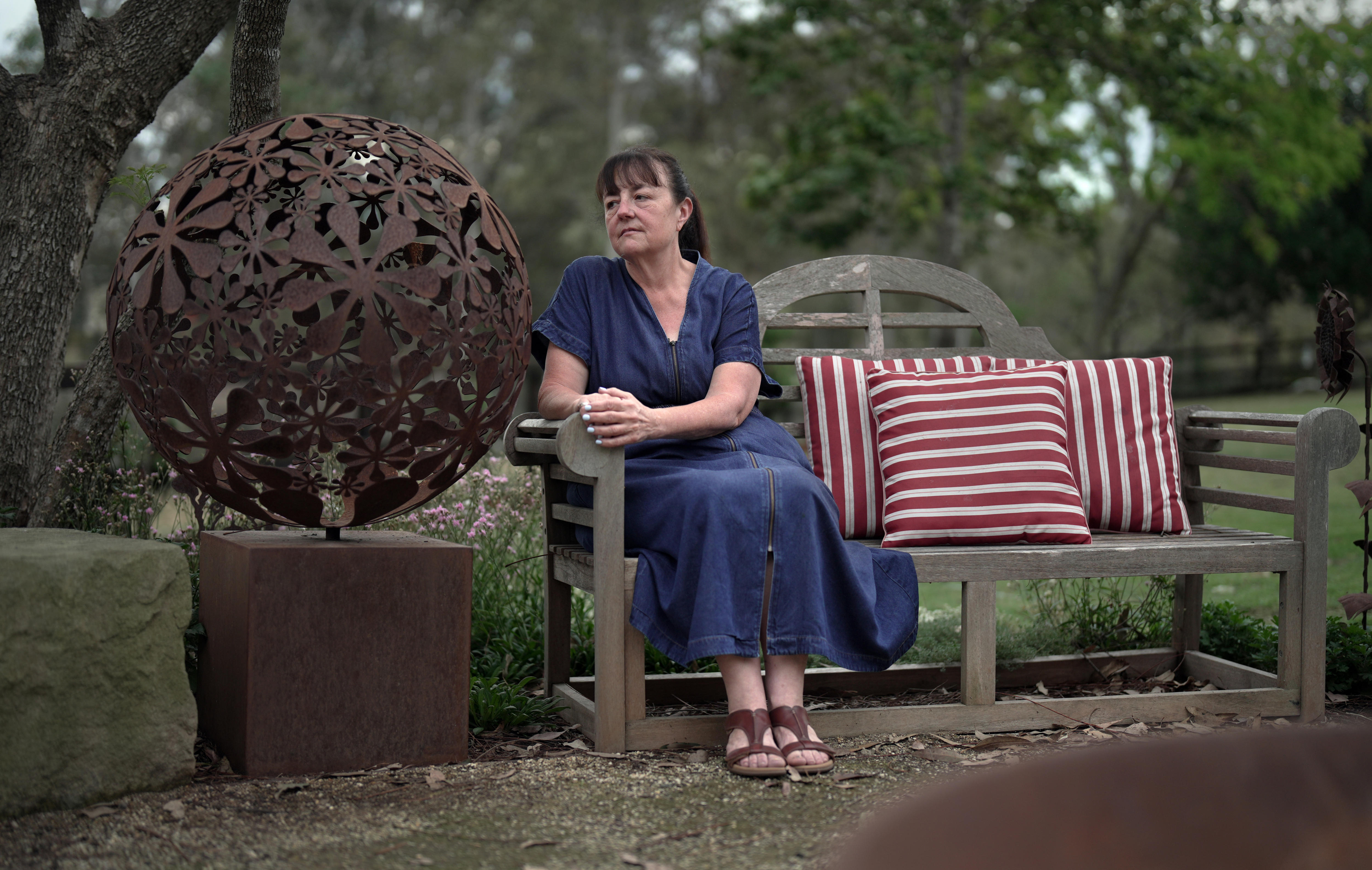 Woman in a navy skirt and top sits on a bench in a garden. A large metal sphere sculpture is to her right.