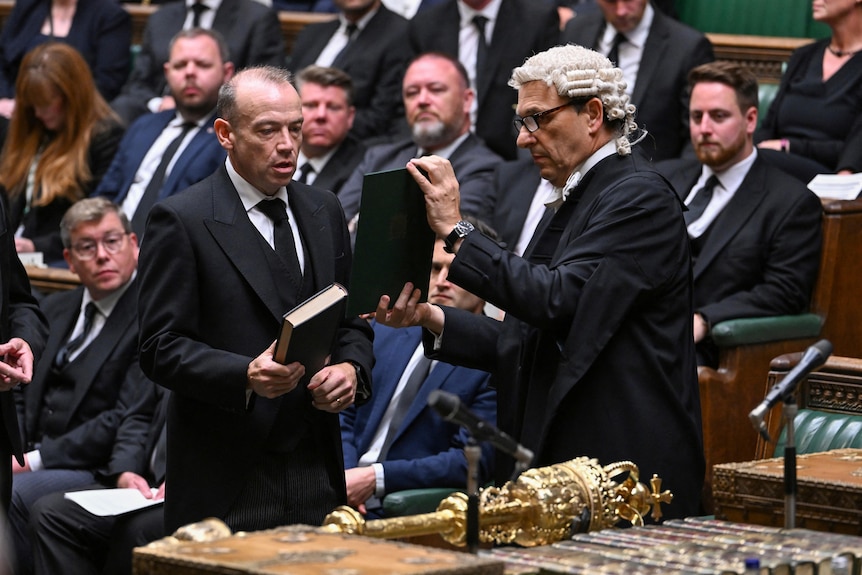Chris Heaton-Harris holds a bible and reads off a page held up by a man in a wig in a parliamentary chamber