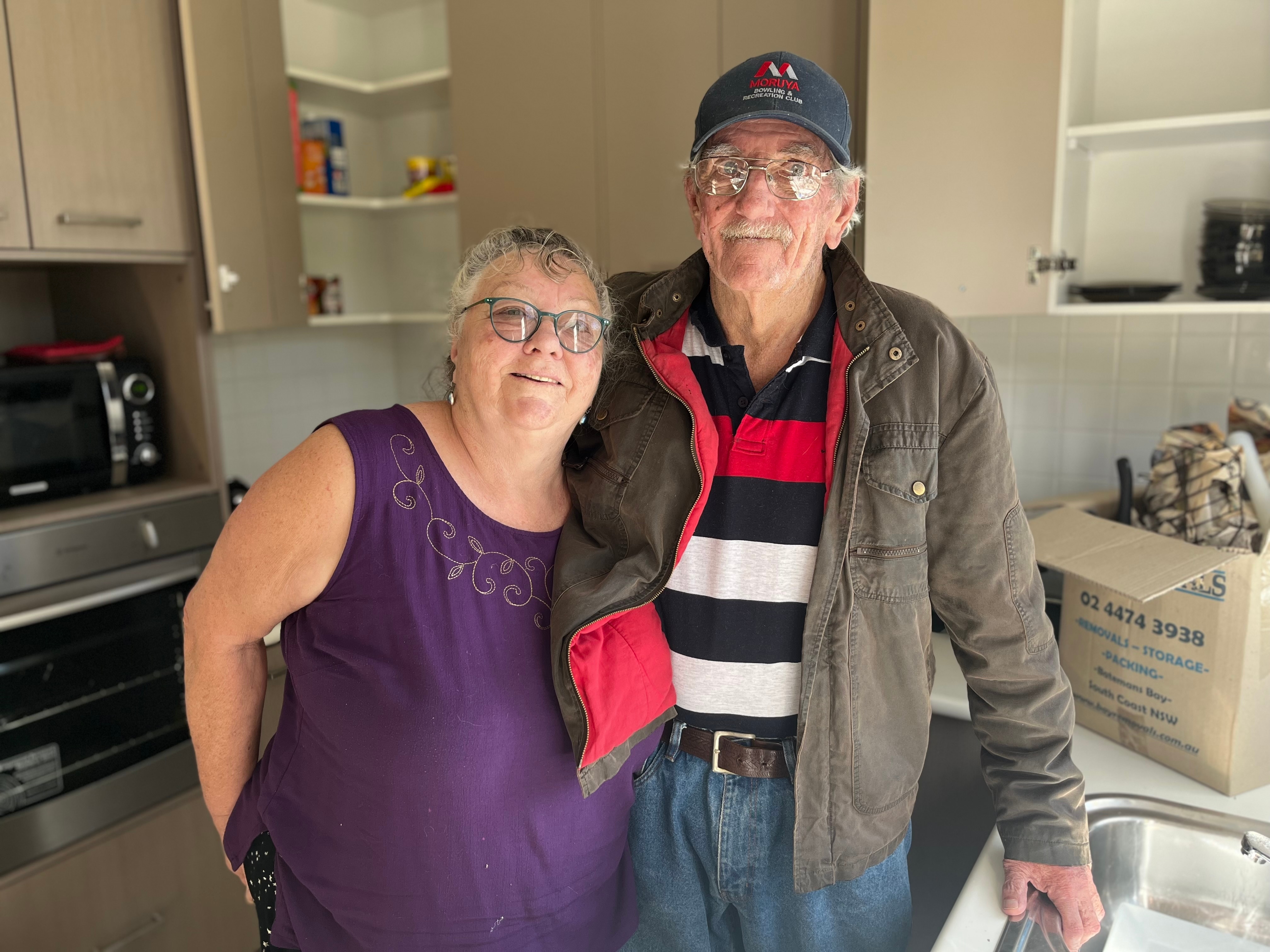 A brother and sister in their 70s stand in a kitchen among moving boxes. 
