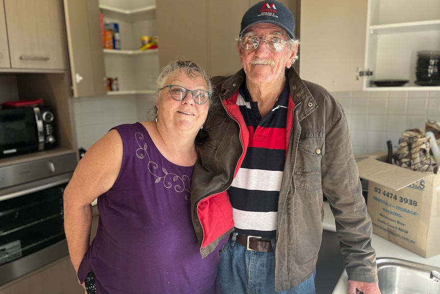 A brother and sister in their 70s stand in a kitchen among moving boxes. 