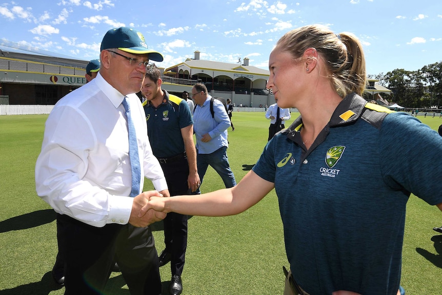 A woman wearing a cricket shirt and cap shake hands with a middle-aged man wearing a business suit.