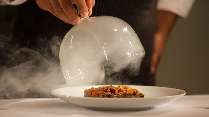Photo of a dish being served, there is a white table cloth and a glass dome with mist over the plate 