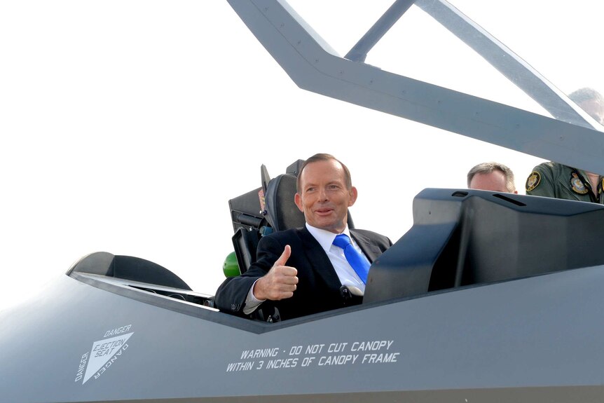 Tony Abbott is smiling and giving a thumbs up from the cabin of a F-35 jet, he is wearing a suit and tie.