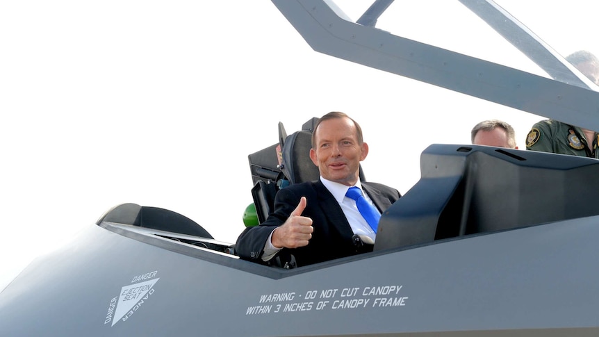 Tony Abbott is smiling and giving a thumbs up from the cabin of a F-35 jet, he is wearing a suit and tie.