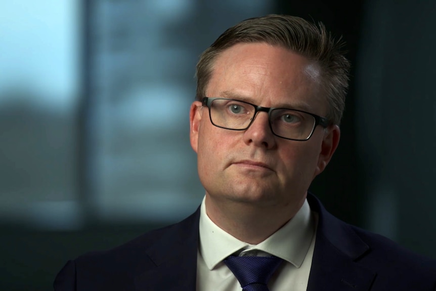 A man in a suit and wearing glasses sitting indoors, with a blank expression. He is looking just to the left of the camera.