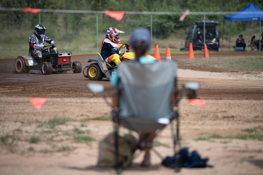 People wearing helmets race ride-on lawn mowers in front of a spectator sitting in a camping chair.
