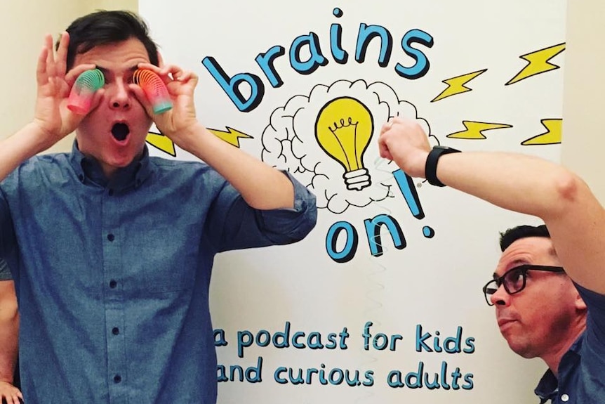 One man holds rainbow slinkies to his eyes while another stretches one out in front of a "Brains on!" poster.