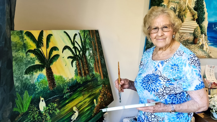 An older woman in a bright outfit paints a tropical scene on a canvas.